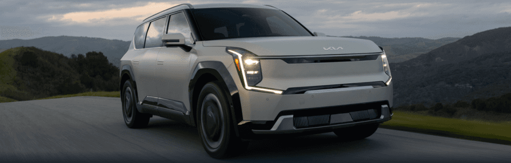 A sleek, white SUV drives on a scenic road with rolling hills and a cloudy sky in the background. The vehicle's design is modern and streamlined.